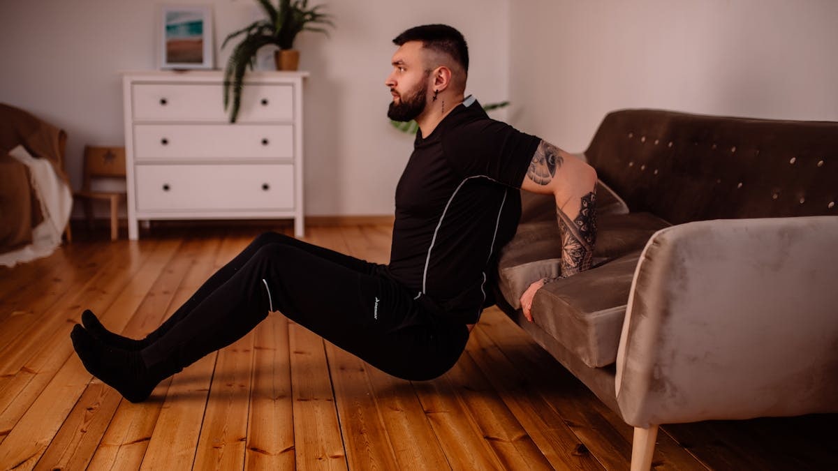 A bearded man in black fitness attire exercising at home, showcasing arm tattoos and a healthy lifestyle.
