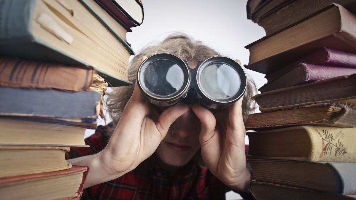 Young unrecognizable person sitting at table with stacked old books and looking through binoculars